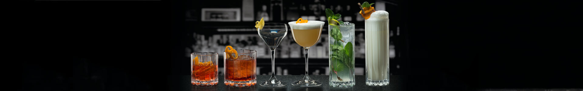 Verschiedene Bargläser aus der Serie Riedel Bar stehen gefüllt mit Cocktails nebeneinander.